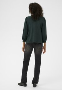Donkergroene, gestructureerde blouse met poffende mouwen, gecombineerd met zwarte recht toe recht aan jeans. Voeten in zwarte schoenen, gezien van achteren.