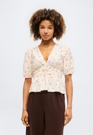 Γυναίκα με σγουρά μαλλιά που φοράει μια κρεμ μπλούζα με ροζ λουλουδάτο σχέδιο και φουσκωτά μανίκια, σε συνδυασμό με σκούρο καφέ παντελόνι, στέκεται μπροστά σε λευκό φόντο.