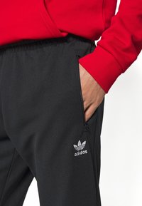 Pantaloni jogger neri realizzati in tessuto morbido con tasche laterali con zip, con il logo bianco Adidas sulla coscia sinistra e il polsino di una felpa rossa in vista.