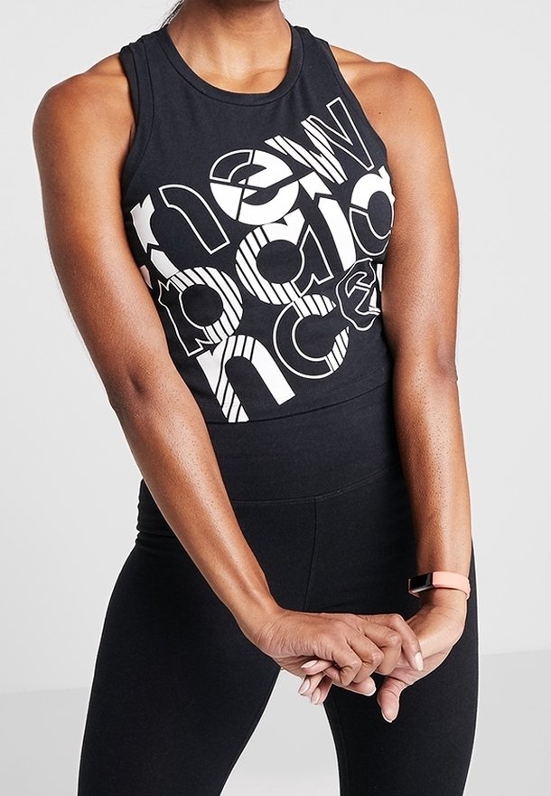 Femme portant un crop top noir avec un texte abstrait blanc en gras, des leggings noirs taille haute, et un bracelet de fitness rose au poignet gauche.