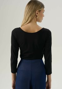 Vrouw met lang blond haar, die gouden oorbellen met een ring draagt, een zwart langemouwen top met een wijde halslijn, en hoog-getailleerde blauwe broek, met de rug naar de kijker gekeerd.