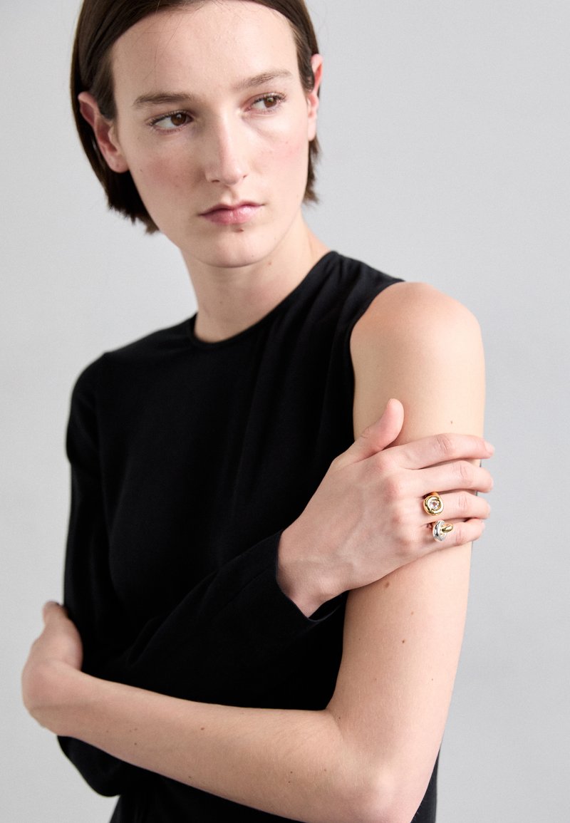 Black long-sleeve top worn by a person, with two rings—one gold and one silver—featuring round settings on the fingers. Light background.