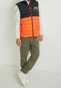 Kamizelka pikowana z czarną górną połową i pomarańczową dolną połową, z zamkiem z przodu i logo "Unlimited Potential". Połączona z oliwkowymi spodniami typu cargo.