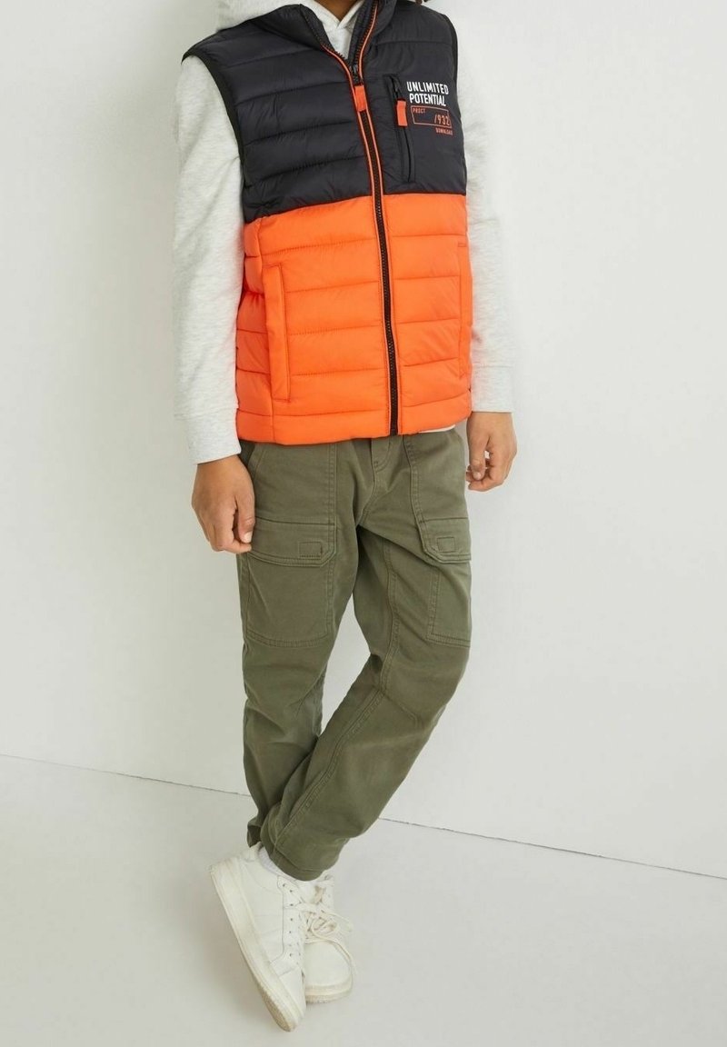 Kamizelka pikowana z czarną górną połową i pomarańczową dolną połową, z zamkiem z przodu i logo "Unlimited Potential". Połączona z oliwkowymi spodniami typu cargo.