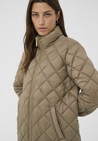 Frau mit langen braunen Haaren, die Ohrringe mit Creolen trägt und eine beigefarbene gesteppte Jacke mit hohem Kragen und Reißverschluss an hat, die Hände hinter dem Rücken.