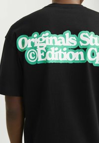 Camiseta de algodón negra con un gráfico de texto en blanco y teal en la parte posterior que dice "Originals Studio © Edition". Manga corta y diseño sencillo.