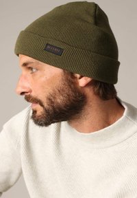 Bonnet tricoté en vert olive avec un revers, arborant une petite étiquette noire avec "DEELUXE" en texte marron. Texture lisse et design ajusté.