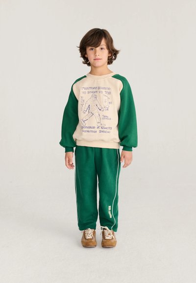 Jeune garçon aux cheveux bruns, debout, portant un sweat-shirt beige et vert avec un graphique de Yéti, un pantalon de survêtement vert assorti et des chaussures marron.