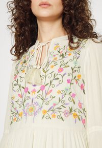 Broderad blus i krämfärg med färgglada blommönster. Har rund halsringning, kvastband och textil med struktur och veck.