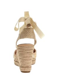 Zapato de esparto beige tejido con superficie texturizada, talón abierto y lazo de tela en el tobillo. Presenta una punta redonda y detalles con patrón.