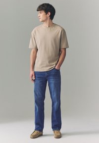 Beiges Baumwoll-T-Shirt mit kurzen Ärmeln, kombiniert mit blauen Jeans aus Denim und braunen Wildlederschuhen. Das Model steht vor einem neutralen Hintergrund.
