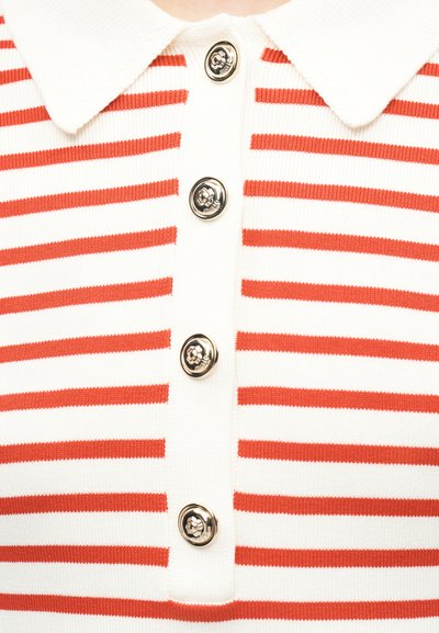 Λευκό πουκάμισο με γιακά και οριζόντιες κόκκινες ρίγες, με τέσσερα μεταλλικά κουμπιά σε σχήμα λουλουδιού κατά μήκος του μπροστινού μέρους.