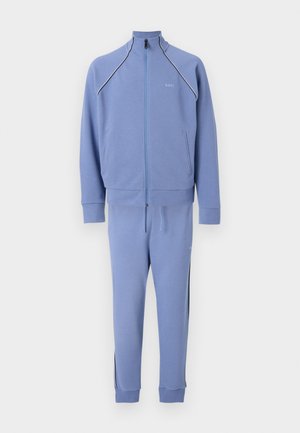 Svetlomodrá športová bunda na zips a zodpovedajúce tepláky s čiernym a bielym lemovaním na pleciach a bokoch nohavíc, zobrazené na bielom pozadí.