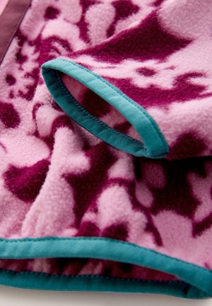 Fleecedecke in Pink mit violetten abstrakten Mustern. Die Kanten sind mit einem türkisen Band eingefasst. Weiche Textur, eine Falte zeigt das geschichtete Design.