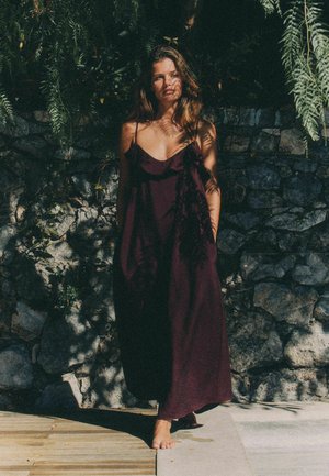 Femeie într-o rochie fără mâneci de culoare mov închis stă desculță pe o pavăză de piatră, cu umbre de frunze proiectate peste ea și un zid de piatră în spate.