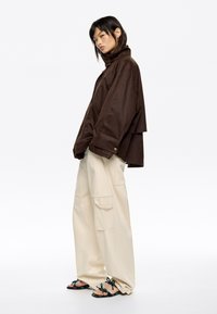 Chaqueta marrón de gran tamaño con cuello alto y detalles en botones, combinada con pantalones cargo beige holgados y sandalias multicolores. Fondo liso.
