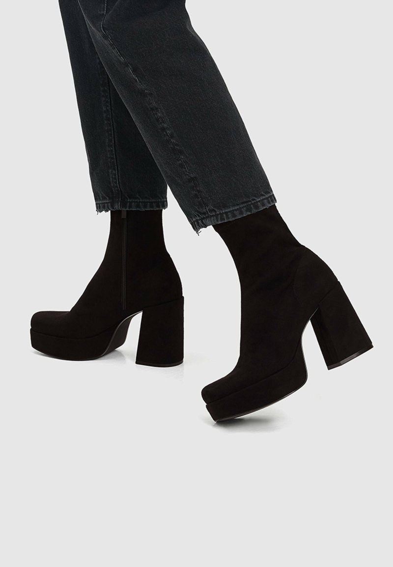 Personne portant des bottines noires à plateforme avec talons blocs et un jean court foncé sur un fond clair uni.