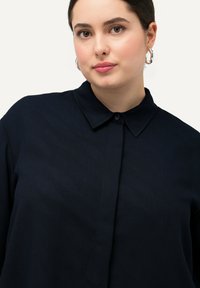 Camicia blu navy con bottoni, realizzata in un tessuto morbido e testurizzato, presenta un colletto classico e una leggera lucentezza, indossata da un modello su uno sfondo chiaro.