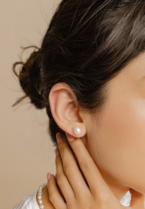 Orecchini a bottoncino in perla bianca su un orecchio di una modella, con una superficie liscia. La modella ha i capelli scuri acconciati in un bun e indossa una collana di perline.