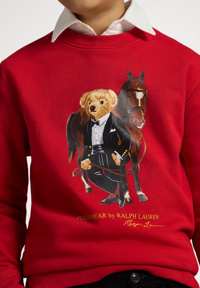 Sweatshirt vermelha com um urso de fraque segurando uma fusta, com um cavalo castanho atrás. Texto impresso: "POLO BEAR by RALPH LAUREN."