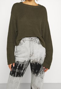 Olivgrüner, gestrickter Pullover mit weitem Ausschnitt und lockerem Sitz, kombiniert mit grauen Batik-Jeans mit schwarz-weißem Muster.