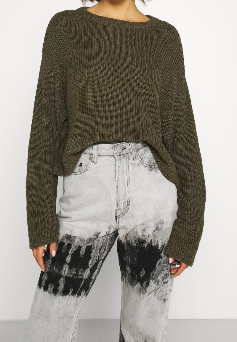 Olivgrüner, gestrickter Pullover mit weitem Ausschnitt und lockerem Sitz, kombiniert mit grauen Batik-Jeans mit schwarz-weißem Muster.