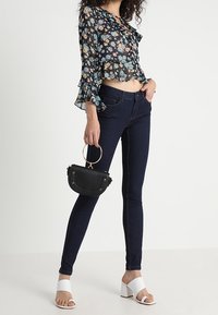 Blusa negra translúcida con estampado floral, con escote con volantes y mangas acampanadas. Jeans ajustados de mezclilla oscuros. Sandalias de tacón bloque blanco. Peque�ño bolso negro con asa en forma de anillo.