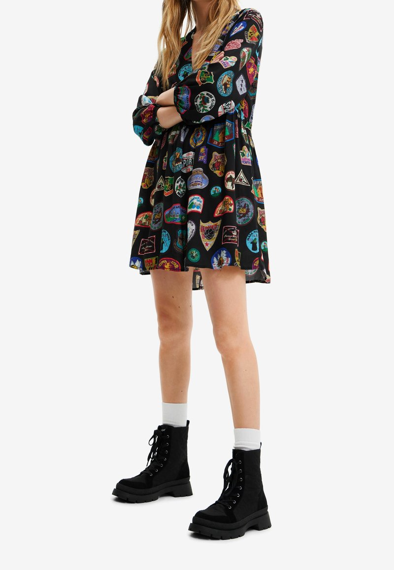 Femme portant une robe noire à manches longues avec des patchs colorés et des bottes noires à lacets avec des chaussettes blanches, debout les bras croisés.