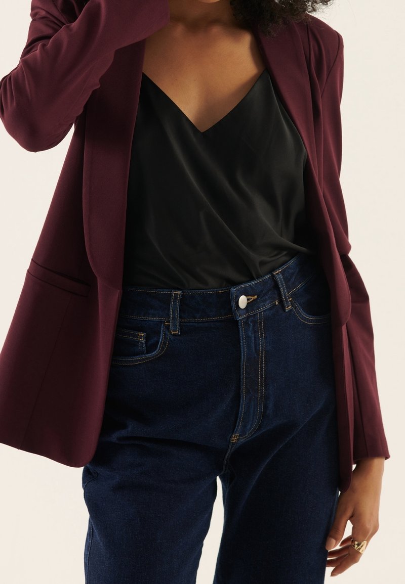 Femme portant un haut noir à col en V, jean taille haute bleu foncé et blazer bordeaux foncé, la main près du visage.