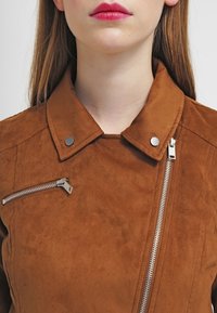 Veste en daim marron avec un col structuré, des accessoires en métal argenté et une fermeture à glissière asymétrique. Comprend une poche poitrine zippée.