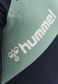 Hummel Sportshirt in Grün- und Marineblau mit glatter Textur und einem großen weißen Logo sowie einer Biene-Grafik auf der Vorderseite.