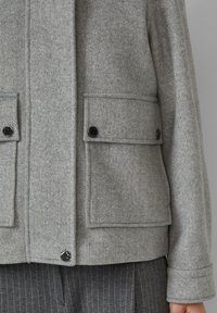 Grau karierten Mantel mit Taschenklappen, schwarzen Knöpfen und einem strukturierten Design, kombiniert mit grau gestreiften Anzughosen.