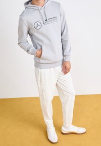Grå hoodie med dragsko, med en logotyp och text. Kombinerad med vita tapered byxor, sidodragkedjor, och vita sneakers med orange detaljer.