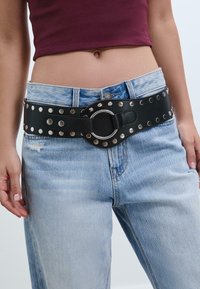 Zwarte leren riem met zilveren studs en een ronde gesp, gedragen over lichtblauwe versleten denim jeans. Gladde textuur, brede ontwerp.