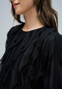 Top nero caratterizzato da una texture a coste con dettagli ondulati a strati lungo le spalle. Collo rotondo e tessuto morbido e leggero.