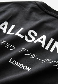 Juodas audinys marškinėliai su drąsiu baltu tekstu, kuriame užrašyta "VISI ŠVENTIEJI" ir "LONDONAS", su papildomais simboliais japonų kalba.