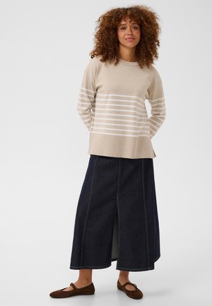 Vrouw met krullend haar die een beige gestreepte trui, donkere lange rok met voorsplit en bruine platte schoenen draagt, staand tegen een effen achtergrond.