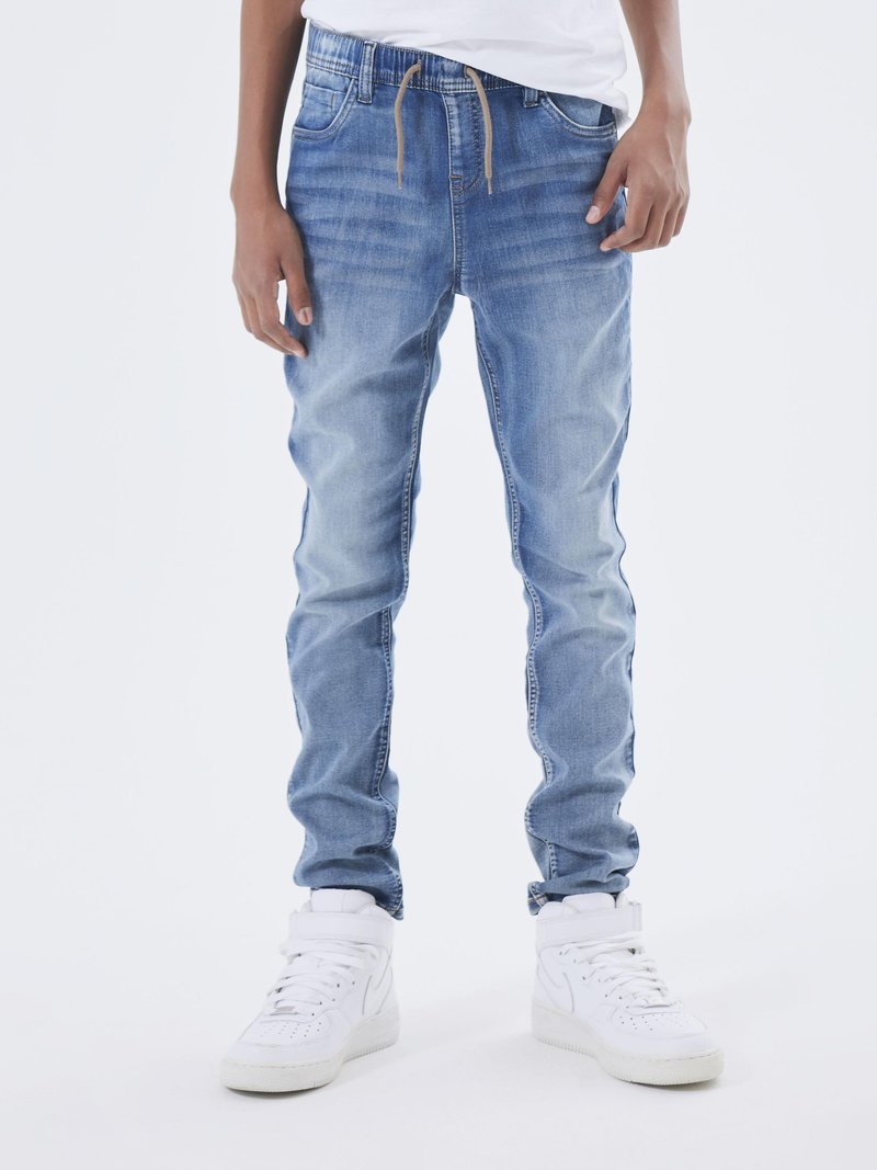 Jeans in denim azzurro chiaro con una vestibilità slim, vita con fondo elasticizzato e due tasche frontali. Indossati con sneakers bianche alte.