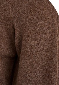 Nærbillede af en brun tekstureret ulden sweater, der viser stoffets bløde, fnuggede strikkemønster og skuldersømsdetalje.