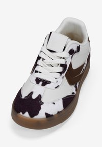 Sneakers bianchi con un motivo a macchie di mucca marroni, con una parte superiore testurizzata, lacci bianchi e un accento laterale marrone. Suola in gomma con grip.