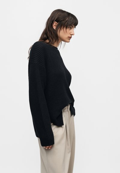 Γυναίκα με μαύρο, φαρδύ πλεκτό πουλόβερ και μπεζ φαρδιά παντελόνια, σε προφίλ, μπροστά από απλό γκρι φόντο.