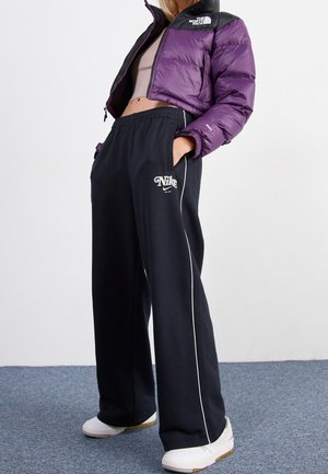 Osoba ubrana w krótka fioletową kurtkę North Face, czarne spodnie dresowe Nike z białym paskiem i białe buty sportowe na szarym dywanie.