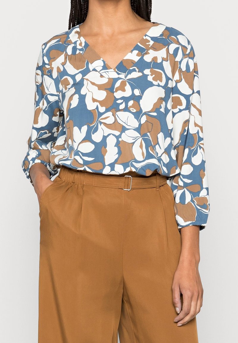 Femme portant un chemisier à fleurs bleu, blanc et marron avec un décolleté en V et des manches trois-quarts, rentrée dans un pantalon marron taille haute avec ceinture.