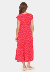 Robe maxi à motifs rouges et roses avec des manches courtes et des épaules dénudées, silhouette fluide, longueur aux genoux, portée avec des ballerines tissées de couleur claire.