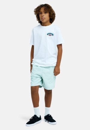 T-shirt en coton blanc avec un graphique coloré, associé à un short vert menthe. Des baskets noires et des chaussettes blanches complètent la tenue.