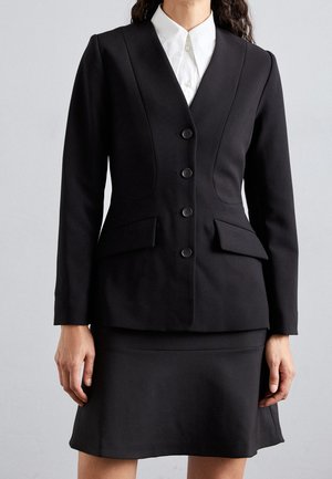 Mujer vistiendo blazer negro entallado con botones sobre camisa blanca con cuello y falda negra a juego sobre fondo liso.