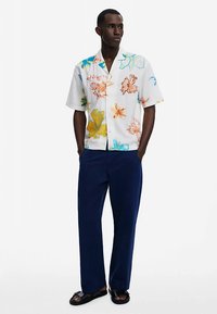 Košulja kratkih rukava na kopčanje s bijelom pozadinom, ukrašena višebojnim cvjetnim uzorcima; uparena s tamnoplavim hlačama i sandalama.