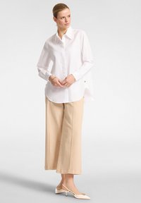 Camicia bianca button-up con maniche lunghe, orlo curvo e dettagli in hardware tono oro, abbinata a pantaloni beige a gamba larga e scarpe slingback color crema.