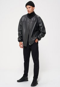 Czarna skórzana kurtka bombowa o gładkiej fakturze, z wysokim kołnierzem, zamkiem z przodu i subtelnymi detalami szwów. Połączona z czarnymi spodniami i sneakersami.