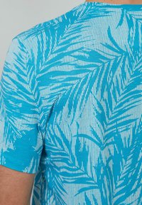 Turquoise t-shirt met een bladerenpatroon in het wit. Korte mouwen, ronde halslijn, de stof heeft een gladde textuur en verticale lijnen.